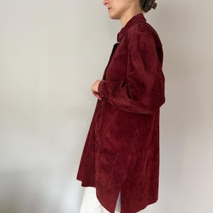 May include: A long, burgundy suede shirt-jacket with a collared neckline and long sleeves. The jacket has two front pockets and a split hem. The garment is knee-length and appears to be worn open.