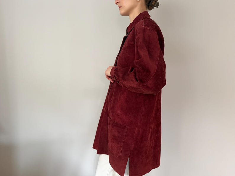 May include: A long, burgundy suede shirt-jacket with a collared neckline and long sleeves. The jacket has two front pockets and a split hem. The garment is knee-length and appears to be worn open.