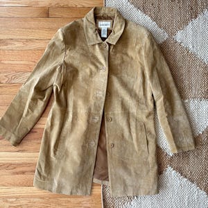 May include: A tan suede jacket with a collared neckline and button-down closure. The jacket has long sleeves and a tailored fit. The garment is displayed on a wooden surface, with a patterned rug visible in the background.