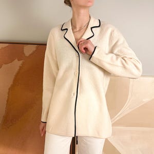 May include: Cream-colored cardigan with black trim. The long-sleeved jacket has a button-up front and a classic collar. The black trim outlines the collar, front, and cuffs. The cardigan is paired with white pants.