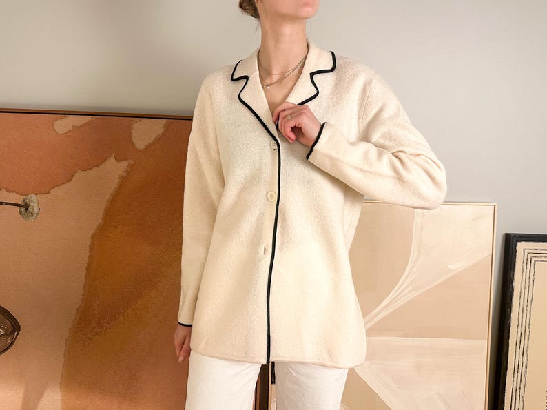 May include: Cream-colored cardigan with black trim. The long-sleeved jacket has a button-up front and a classic collar. The black trim outlines the collar, front, and cuffs. The cardigan is paired with white pants.