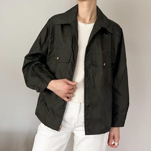 May include: Dark olive green jacket with a collared neckline, two chest pockets, and a zip-up front. The jacket has gold-colored buttons and is worn over a white top and white pants. The jacket's sleeves are long.