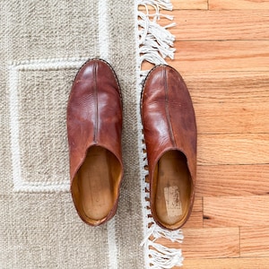 May include: A pair of brown leather slippers with a rounded toe, set on a beige rug with white trim and a wooden floor. The slippers have a worn appearance, suggesting comfort and use.