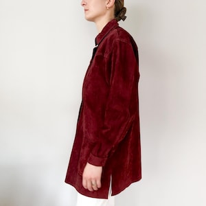 May include: A burgundy-colored button-down shirt with long sleeves. The shirt has a collared neckline and a relaxed fit. The fabric appears to be a soft, suede-like material. The shirt is worn with white pants.