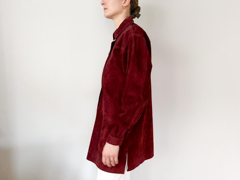 May include: A burgundy-colored button-down shirt with long sleeves. The shirt has a collared neckline and a relaxed fit. The fabric appears to be a soft, suede-like material. The shirt is worn with white pants.