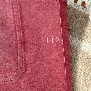 May include: Close-up of a red leather item with the number "112" embroidered in white. The item has a stitched border and a textured surface. The background includes a beige and white woven textile.