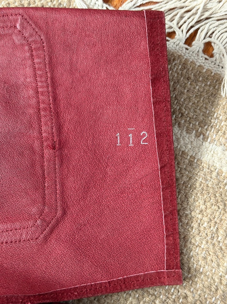 May include: Close-up of a red leather item with the number "112" embroidered in white. The item has a stitched border and a textured surface. The background includes a beige and white woven textile.