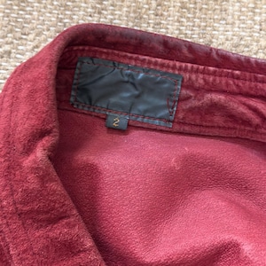 May include: Close-up of a burgundy-colored shirt collar. The shirt has a soft, textured appearance. A black label is sewn into the collar, with the number "2" visible below it. The fabric appears to be slightly worn.
