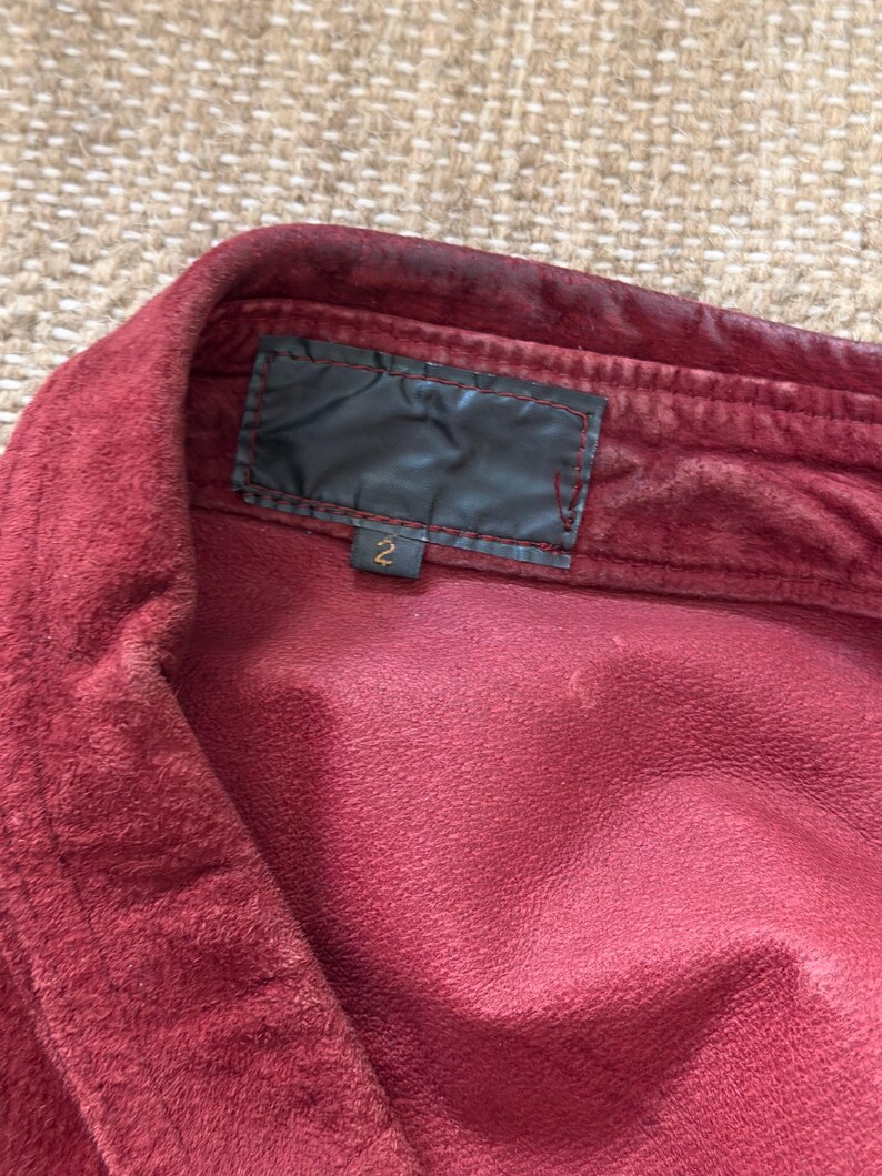 May include: Close-up of a burgundy-colored shirt collar. The shirt has a soft, textured appearance. A black label is sewn into the collar, with the number "2" visible below it. The fabric appears to be slightly worn.
