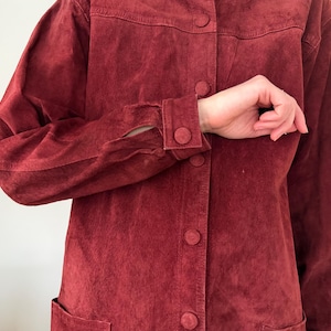 May include: A burgundy suede jacket with a button-up front, two front pockets, and buttoned cuffs. The jacket has a classic design and a soft, textured appearance. The sleeves are long and the jacket appears to be well-worn.