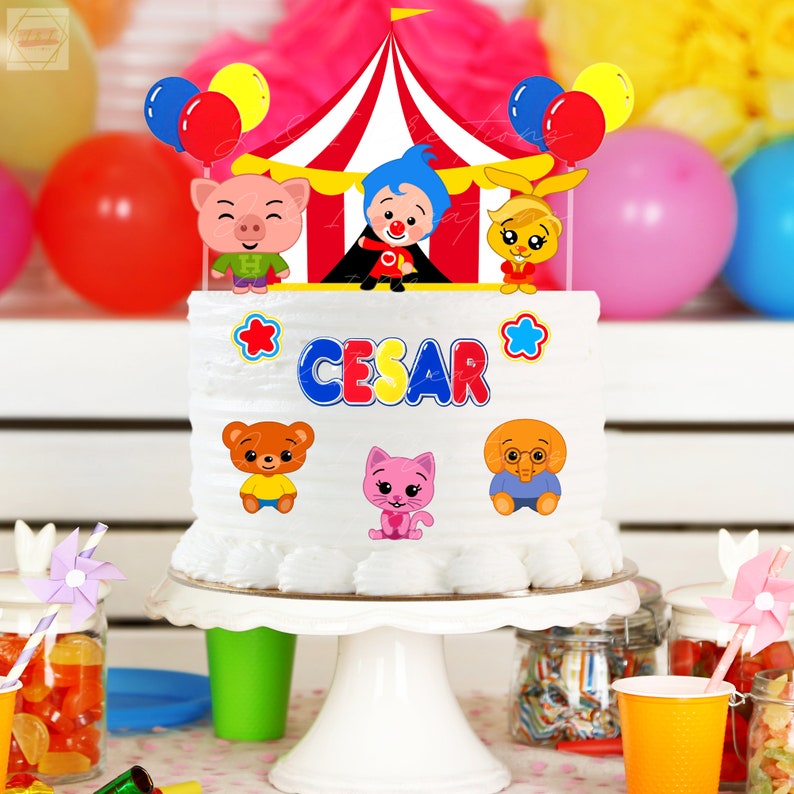 May include: A white cake with a circus theme. The cake is decorated with a red and white striped tent, colorful balloons, and cartoon characters. The cake is topped with the name "CESAR" in colorful letters.