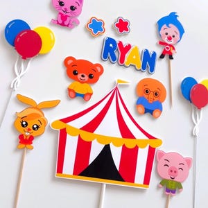 May include: A set of cake toppers featuring characters from the CoComelon cartoon show. The toppers include a red and white striped circus tent, a pink pig, a yellow bunny, a brown bear, a blue elephant, a pink cat, a clown, and two balloons. The name "RYAN" is spelled out in colorful letters.