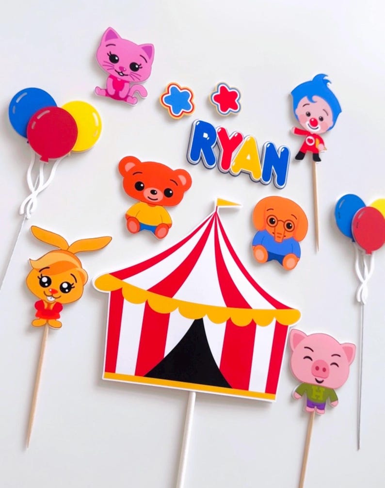 May include: A set of cake toppers featuring characters from the CoComelon cartoon show. The toppers include a red and white striped circus tent, a pink pig, a yellow bunny, a brown bear, a blue elephant, a pink cat, a clown, and two balloons. The name "RYAN" is spelled out in colorful letters.