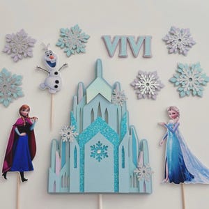 May include: A light blue castle cake topper with glitter accents, surrounded by snowflake decorations and character cutouts of Anna, Elsa, and Olaf. The letters "VIVI" are also included.