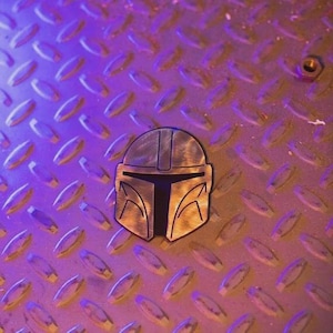 May include: A metallic Mandalorian helmet emblem with a black visor, set against a textured, diamond-plate background. The emblem is a bronze color, and the background has a purple and gold hue.