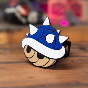 May include: A blue, white, and tan cartoon-style accessory shaped like a spiky shell. The accessory has a black band and is set against a wooden surface. The design is reminiscent of a video game character.