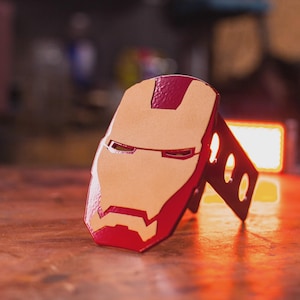 May include: A red and tan Iron Man mask-shaped phone stand. The stand is made of a sturdy material and features the iconic face design with eye and mouth cutouts. The stand is designed to hold a phone upright.
