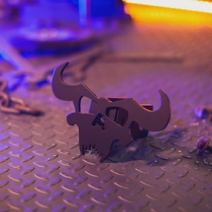 May include: A dark metal skull-shaped hitch cover with curved horns and a detailed skull design. The hitch cover is designed to fit a standard 2-inch receiver. The background is a diamond-plate surface.