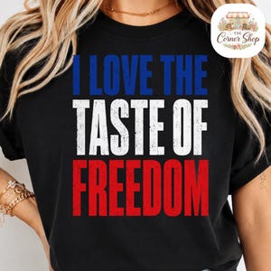 Peut inclure: Un t-shirt noir avec le texte I LOVE THE TASTE OF FREEDOM imprimé dans une police usée. Le mot I LOVE THE est en bleu, TASTE OF est en blanc et FREEDOM est en rouge, créant un schéma de couleurs patriotique sur le devant du vêtement.