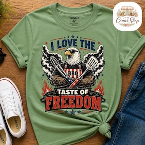 Peut inclure: Un t-shirt vert sauge à col rond avec un motif d'aigle à tête blanche portant des lunettes de soleil et un tablier de barbecue. L'aigle tient des pinces et une spatule. Le texte indique I LOVE THE TASTE OF FREEDOM, avec des flammes et de la fumée.