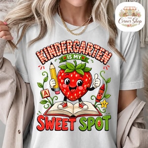 Peut inclure: Un t-shirt blanc avec un motif représentant une fraise souriante debout sur un livre ouvert. La fraise tient un crayon jaune et est entourée d'un crayon rouge, d'un bloc lettre A et du texte "KINDERGARTEN IS MY SWEET SPOT".