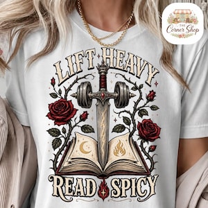 Peut inclure: Un t-shirt blanc avec une impression graphique représentant une épée transperçant un livre ouvert, entouré de roses rouges et d'haltères. Le texte indique LIFT HEAVY en haut et READ SPICY en bas, avec une lune et une flamme sur les pages.