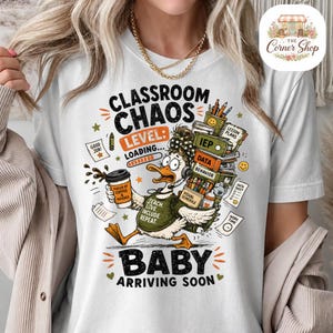 Peut inclure: Un t-shirt blanc avec un motif de canard portant un casque et un haut vert indiquant Teach, Love, Include, Repeat. Le texte affiche Classroom Chaos Level: Loading... Baby Arriving Soon, accompagné d'illustrations de café, livres et fournitures.