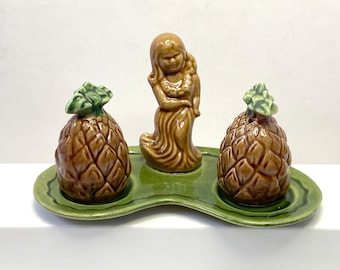 Pode incluir: Uma bandeja de servir de cerâmica com um glacê verde apresenta três estatuetas de cerâmica. Uma estatueta de uma dançarina hula está no centro, e dois saleiros e pimenteiros em forma de abacaxi estão de cada lado.
