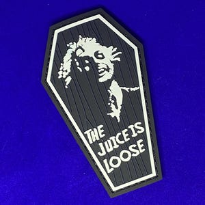 May include: A black and white patch shaped like a coffin with a white graphic of a person's head and the text "The Juice is Loose".