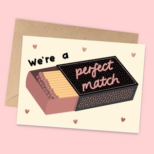 May include: A card with a matchbox illustration and the text "We're a perfect match". The matchbox is open and has a pink and black design with small hearts. The card has a cream background with pink hearts.