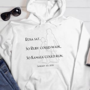May include: White hoodie with a black graphic print of three women walking. The text reads "ROSA SAT. SO RUBY COULD WALK. SO KAMALA COULD RUN. JANUARY 20, 2021".