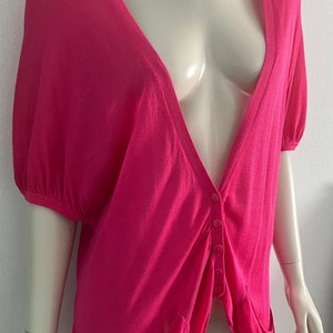Könnte beinhalten: Ein leuchtend pinkfarbener Cardigan mit tiefem V-Ausschnitt und kurzen Ärmeln. Der Cardigan hat einen Knopfverschluss und zwei Taschen.
