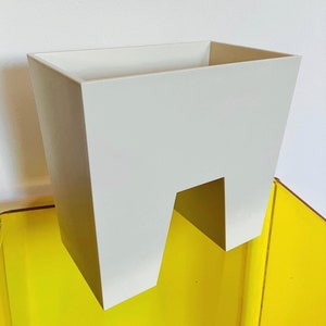 May include: A modern, off-white planter with a unique cut-out design. The square-shaped container has a minimalist aesthetic, with clean lines and a solid color. The planter is set against a bright yellow surface and a white wall.