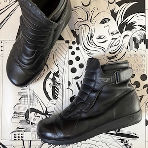 Könnte beinhalten: Ein Paar schwarze Leder-Stiefeletten mit gestepptem Design. Die Stiefeletten haben einen Reißverschluss an der Seite und eine Schnalle oben.