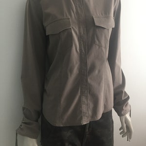 Puede incluir: Una camisa de manga larga de color marrón claro con dos bolsillos de solapa y un detalle de corbata en los puños.