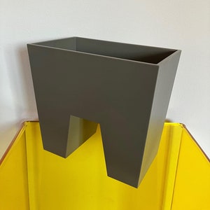 May include: A dark gray planter with a unique design that allows it to be placed over the edge of a shelf or table. The planter has a rectangular shape with a cut-out in the bottom that creates a shelf-like feature.