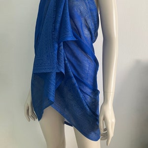Puede incluir: Un pareo azul vibrante con un patrón sutil y repetitivo. La tela ligera está drapeada sobre un maniquí, mostrando su textura fluida. El pareo es un accesorio versátil, perfecto para la playa o como una envoltura elegante.