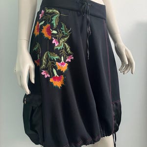 May include: Black skirt with floral embroidery in orange, pink, and green. The skirt has a drawstring waist and a bubble hem. Features a side pocket and pink stitching details. The skirt is made of a soft fabric.