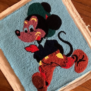 以下が含まれることがあります： 赤い服と黄色の服を着たミッキーマウスが描かれた、ヴィンテージの針仕事による壁掛け。緑色の帽子と黒い耳をしています。背景は薄い青色です。
