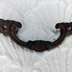 May include: A rusty, cast iron door handle with a decorative, ornate design. The handle is shaped like a D with a curved top and a flat bottom.