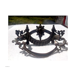 May include: Ornate antique bronze drawer pull with a curved handle and decorative floral and scrollwork details. The hardware features a central floral design and symmetrical side elements, perfect for vintage furniture restoration.