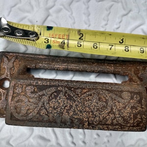 May include: A vintage, ornate, cast iron mail slot with a decorative floral design. The slot is approximately 3.5 inches wide and 1.5 inches tall. The slot is rusty and has a dark brown patina.