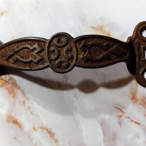 May include: A rusty cast iron door handle with an ornate design. The handle has a keyhole and a decorative center piece.