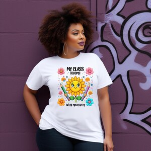 May include: White t-shirt with a colorful graphic featuring a smiling sunflower, pencils, and flowers. The text "MY CLASS BLOOMS WITH CREATIVITY" is printed above and below the sunflower design. The shirt is worn by a person.