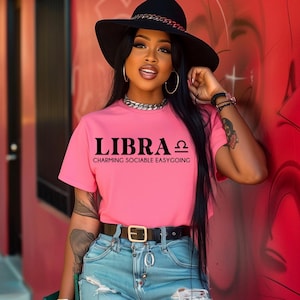 May include: A pink t-shirt with the text "LIBRA ♎ CHARMING SOCIABLE EASYGOING" printed on the front. The model is wearing a black hat and jeans.
