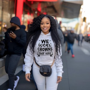 以下が含まれることがあります： 白いパーカーとそれに合わせたパンツを着た女性。パーカーには黒字で「WE WEAR CROWNS OVER HERE」と書かれています。彼女はまた、金のチェーンストラップが付いた黒いハンドバッグを持っています。背景は、他の人々がいる街の通りです。