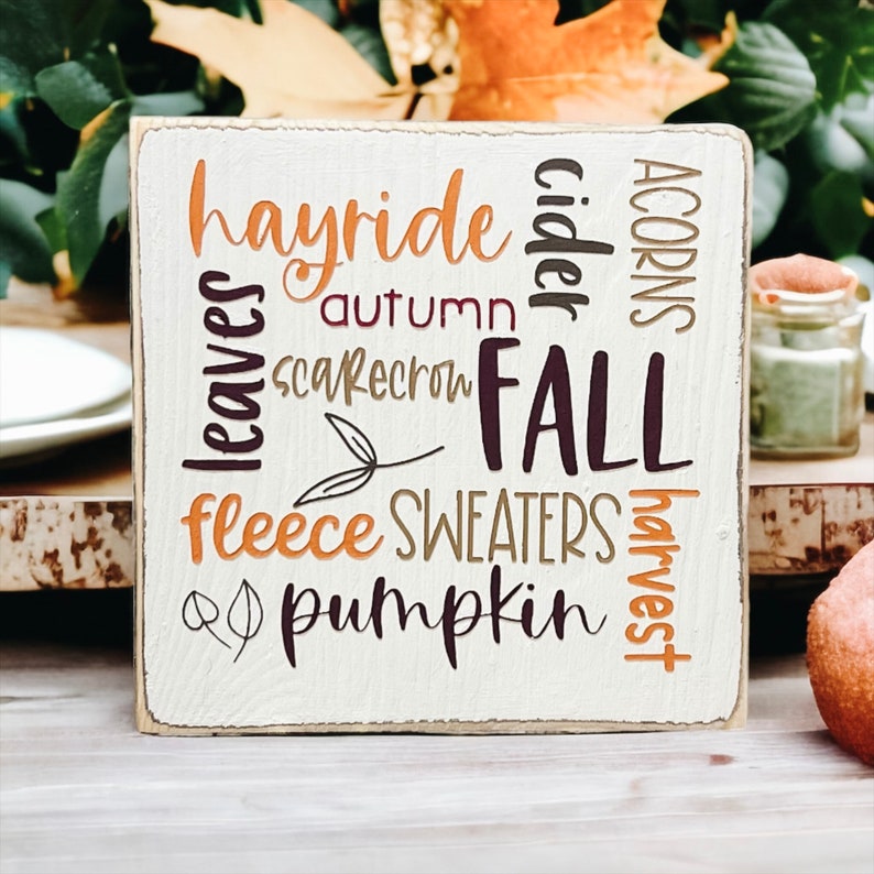 May include: A white wooden sign with the words 'hayride', 'cider', 'acorns', 'leaves', 'autumn', 'scarecrow', 'FALL', 'fleece', 'SWEATERS', 'harvest', and 'pumpkin' written in various colors and fonts.
