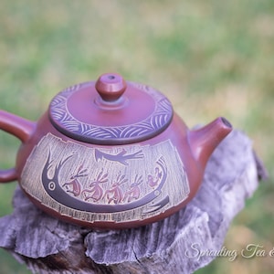 May include: A red clay teapot with a carved design of animals and a bird. The teapot is sitting on a wooden stump.