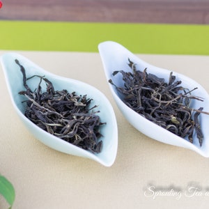 May include: Two white ceramic bowls filled with loose leaf tea. The tea is dark green and brown in color. The bowls are on a light beige surface with a green and white pattern.