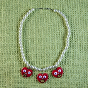 May include: A white pearl necklace with three red heart-shaped pendants. Each pendant has a painted face with blue eyes and a red mouth.
